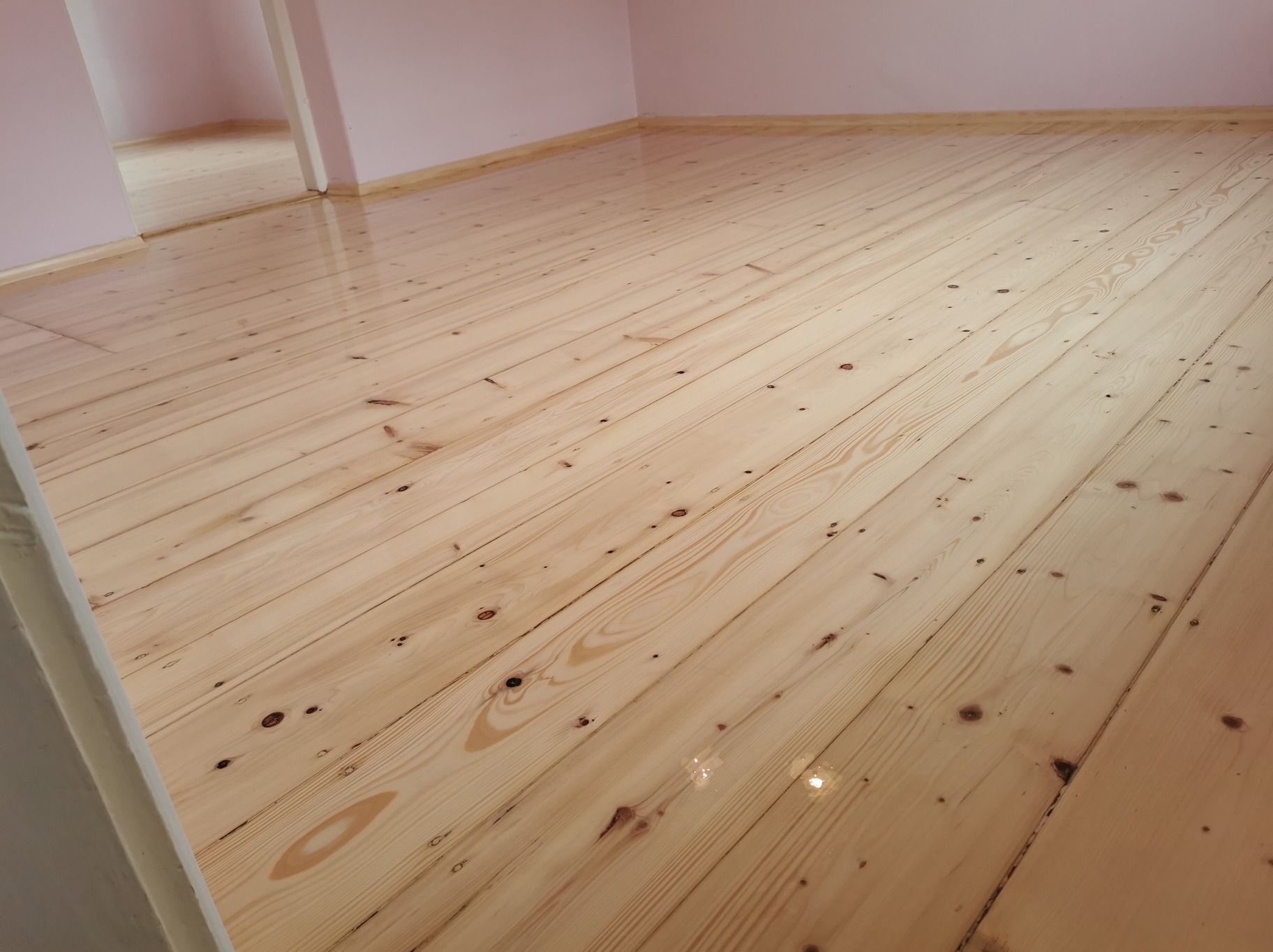 Jasna, lakierowana podłoga z desek w pokoju o różowych ścianach, widok z niskiej perspektywy, ukazujący połysk powierzchni i naturalne usłojenie drewna.