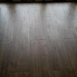 Ułożone ciemnobrązowe panele podłogowe z widocznymi drzwiami balkonowymi i żwirowym podłożem na zewnątrz.