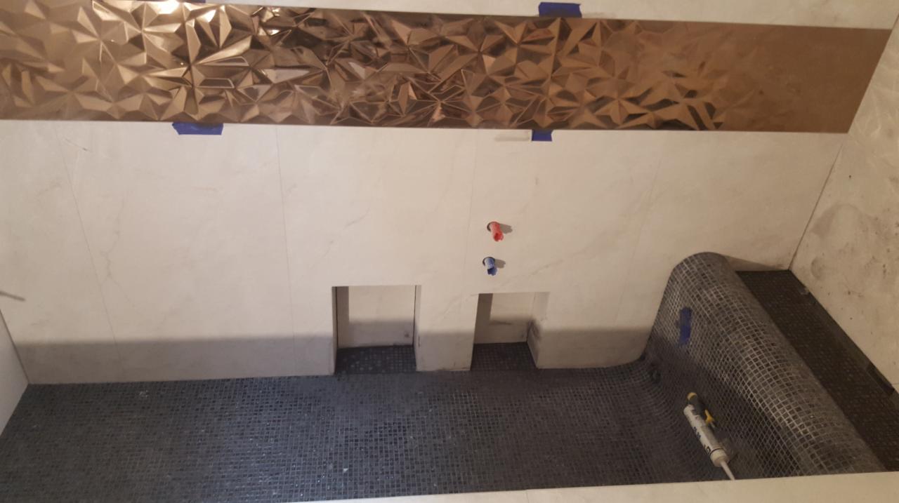 Wnętrze łazienki w trakcie remontu: ułożona mozaika podłogowa, białe płytki ścienne z ozdobnym, metalicznym pasem i wycięciami na półki, widoczna siatka wzmacniająca i rurki instalacji wodnej.