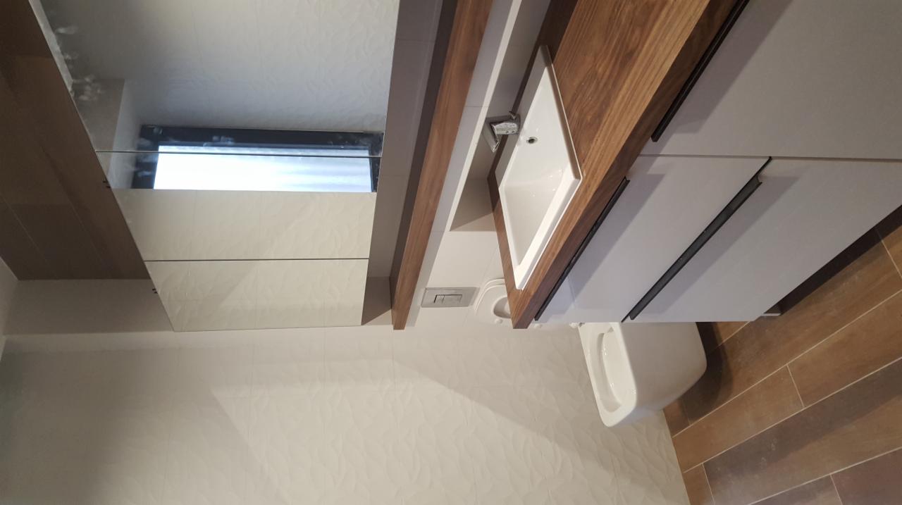 Nowoczesna łazienka z białymi płytkami o subtelnym wzorze, drewnianym blatem pod umywalką i szafkami z ciemnymi uchwytami, oraz wiszącą miską WC.