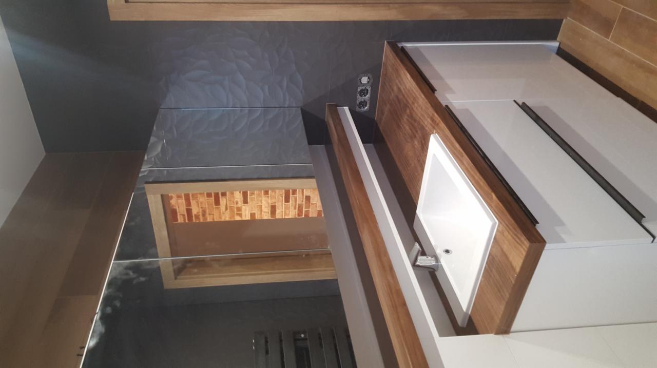 Nowoczesna łazienka z umywalką w drewnianej obudowie, czarnymi płytkami z delikatnym wzorem na ścianie i lustrem odbijającym fragment ściany z cegiełkami.