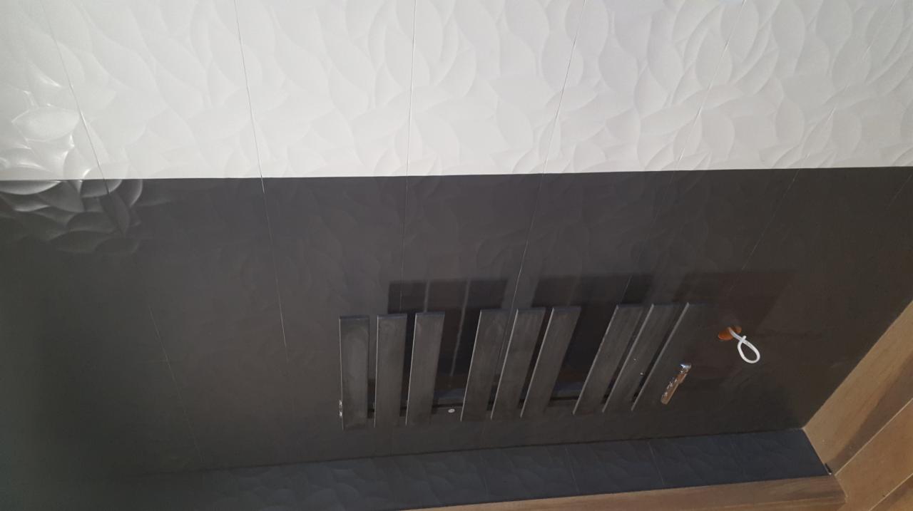 Wykończona ściana z ciemnych, błyszczących płytek z dekoracyjną strukturą, oddzielona od białego sufitu z trójwymiarowym wzorem; widoczne metalowe elementy dekoracyjne i otwór z przewodem.