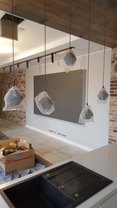 Wnętrze w trakcie prac wykończeniowych: surowa ściana z cegły, szara ściana pomalowana farbą, podwieszane lampy w folii ochronnej, widoczny fragment blatu kuchennego ze zlewem i kartonowe pudło.