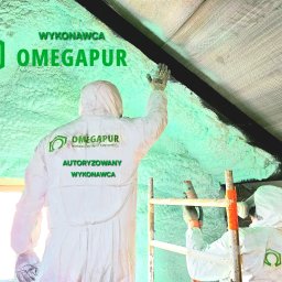 Rehouse MK - Pracownicy w kombinezonach ochronnych aplikują piankę PUR na poddaszu, widoczna struktura pianki i logo firmy wykonawczej OMEGAPUR na kombinezonach.