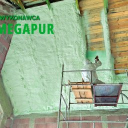 Rehouse MK - Pracownik w kombinezonie ochronnym aplikuje piankę izolacyjną na poddaszu budynku, widoczny rusztowanie i logo firmy wykonawczej.