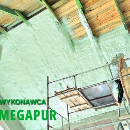 Rehouse MK - Aplikacja izolacji pianką na poddaszu, widoczny pracownik w kombinezonie ochronnym na rusztowaniu, świeżo nałożona piana w jasnym odcieniu zieleni pokrywa krokwie i ściany szczytowe, drewniane...