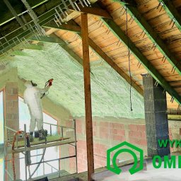 Rehouse MK - Pracownik w kombinezonie ochronnym aplikuje piankę izolacyjną na poddaszu, widoczne krokwie i elementy konstrukcyjne dachu, rusztowanie i logo firmy na pierwszym planie.