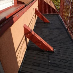 Rehouse MK - Balkon z nową, czarną izolacją przeciwwodną, wsparty na czerwonych, trójkątnych wspornikach przymocowanych do ściany budynku, z widoczną metalową balustradą i elementami dachu w tle.