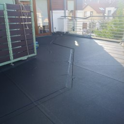 Rehouse MK - Czarna, gumowa izolacja przeciwwodna na tarasie, widoczne łączenia, fragment drewnianej osłony balkonu z zielonymi ramami, stalowa balustrada i budynki w tle.