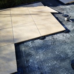 Rehouse MK - Układanie jasnobeżowych płyt na regulowanych wspornikach tarasowych na czarnej hydroizolacji, widoczne ślady robocze i gumowy młotek.