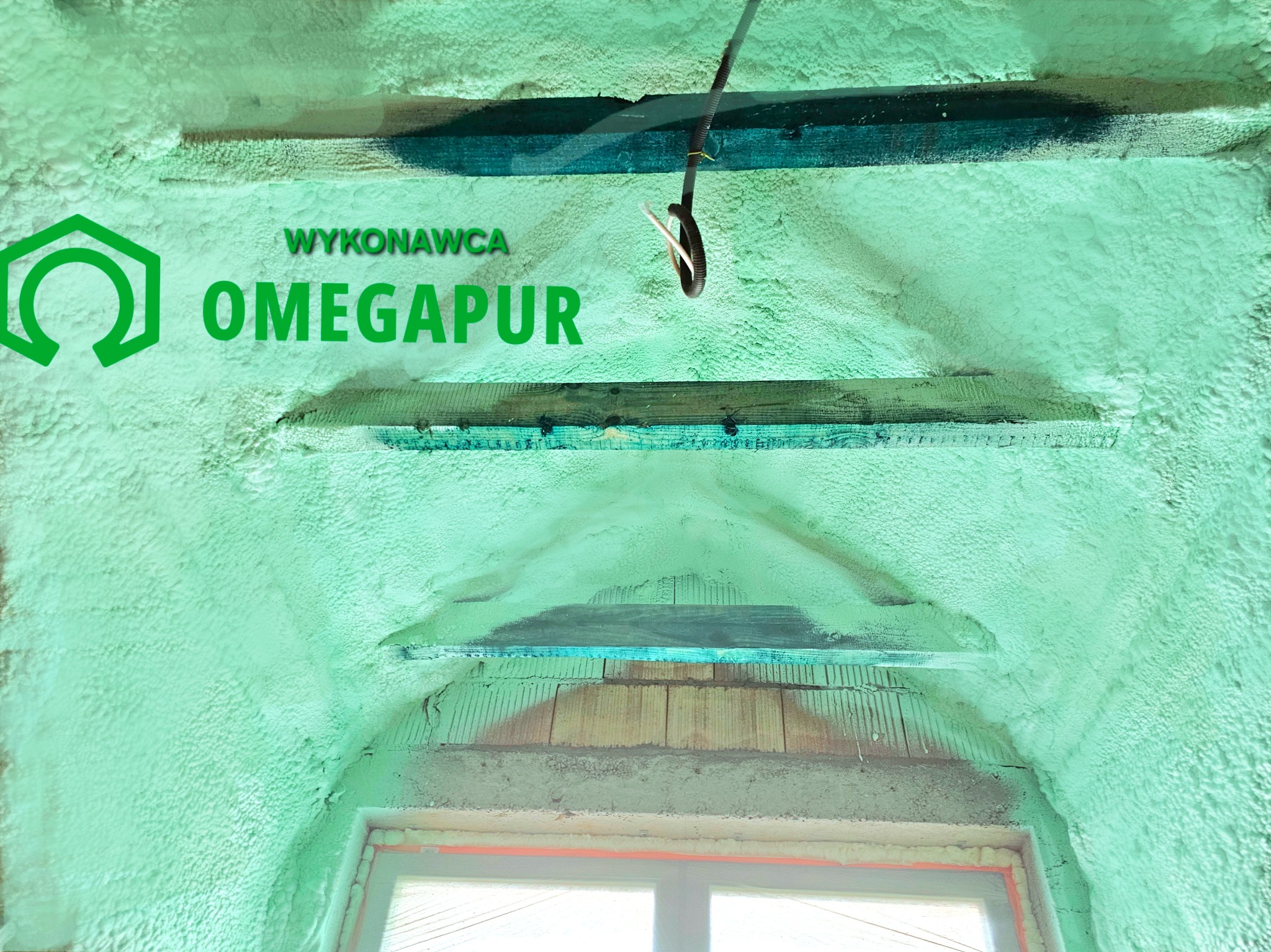 Aplikacja izolacji pianką na poddaszu z widocznym logo wykonawcy Omegapur i elementami drewnianej konstrukcji oraz przewodami elektrycznymi.