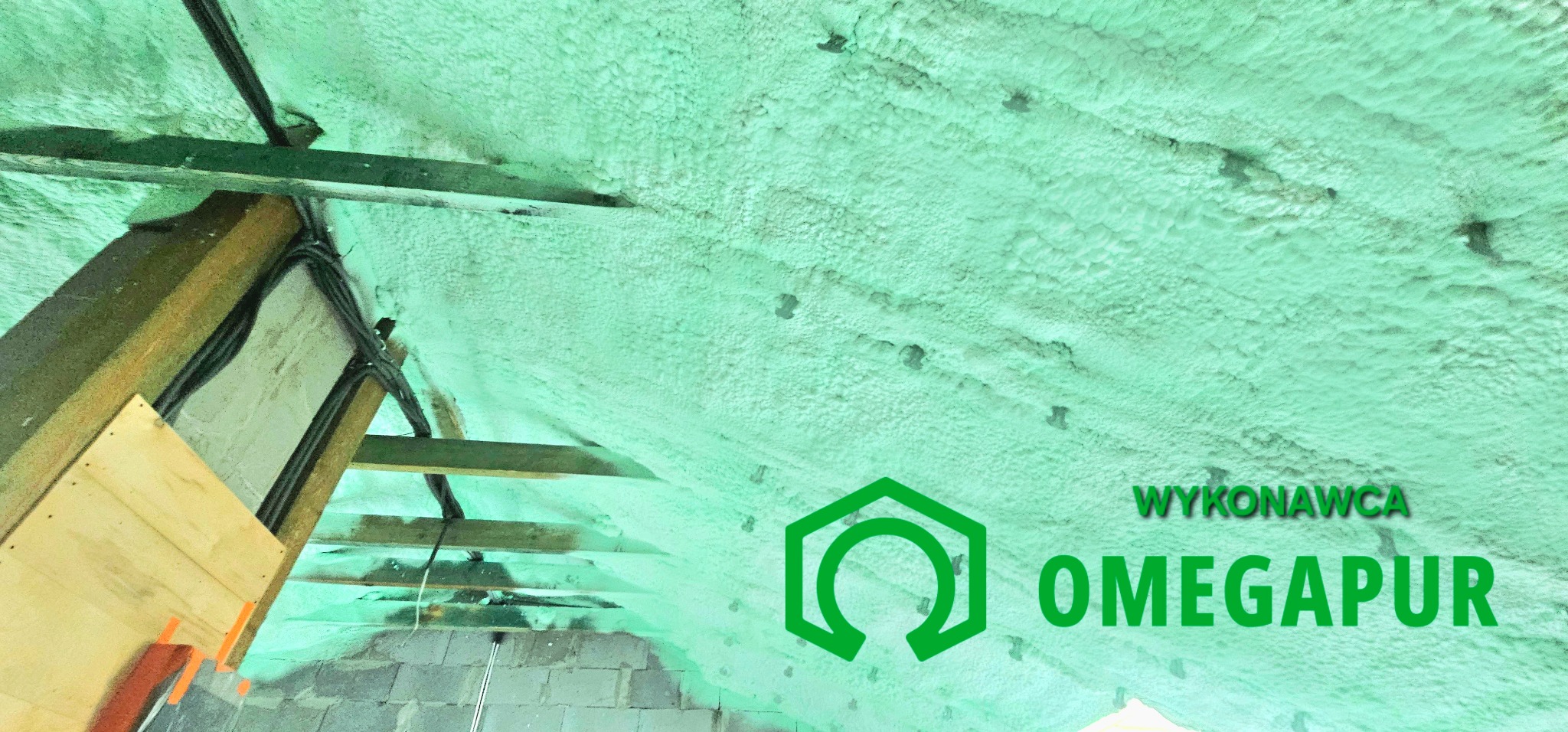 Poddasze z widoczną konstrukcją drewnianą i instalacją elektryczną pokryte warstwą izolacyjnej pianki poliuretanowej, z naniesionym logo firmy wykonawczej OMEGAPUR.