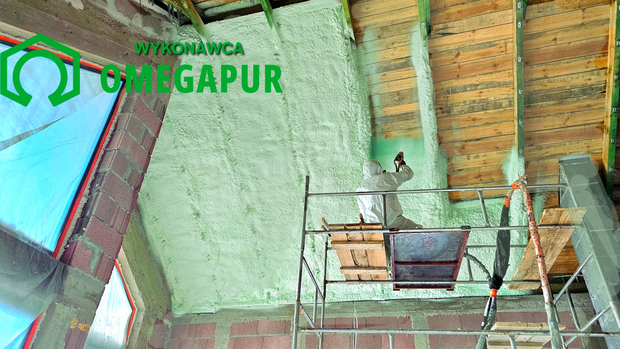 Pracownik w kombinezonie ochronnym aplikuje piankę izolacyjną na poddaszu budynku, widoczny rusztowanie i logo firmy wykonawczej.