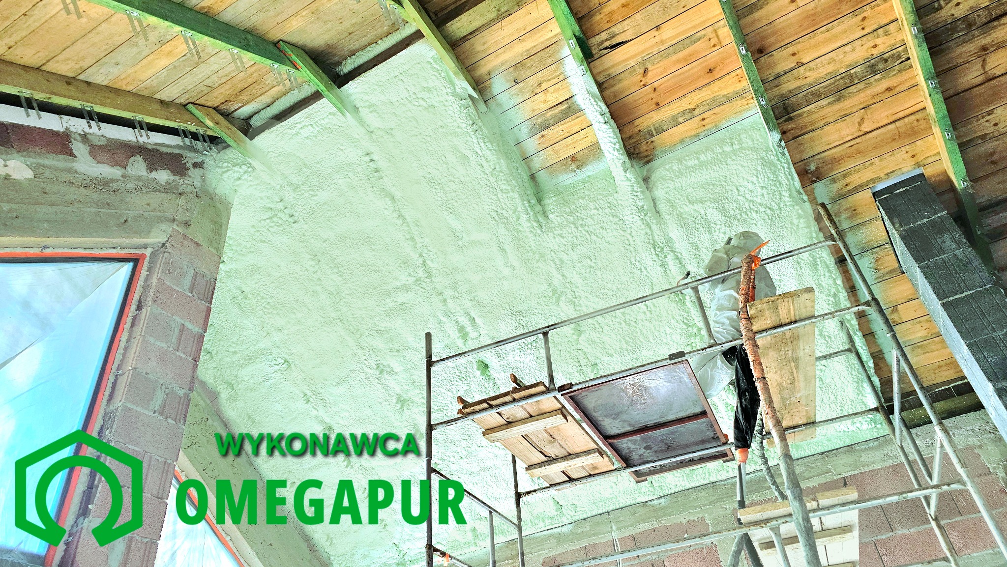 Aplikacja izolacji pianką na poddaszu, widoczny pracownik w kombinezonie ochronnym na rusztowaniu, świeżo nałożona piana w jasnym odcieniu zieleni pokrywa krokwie i ściany szczytowe, drewniane...