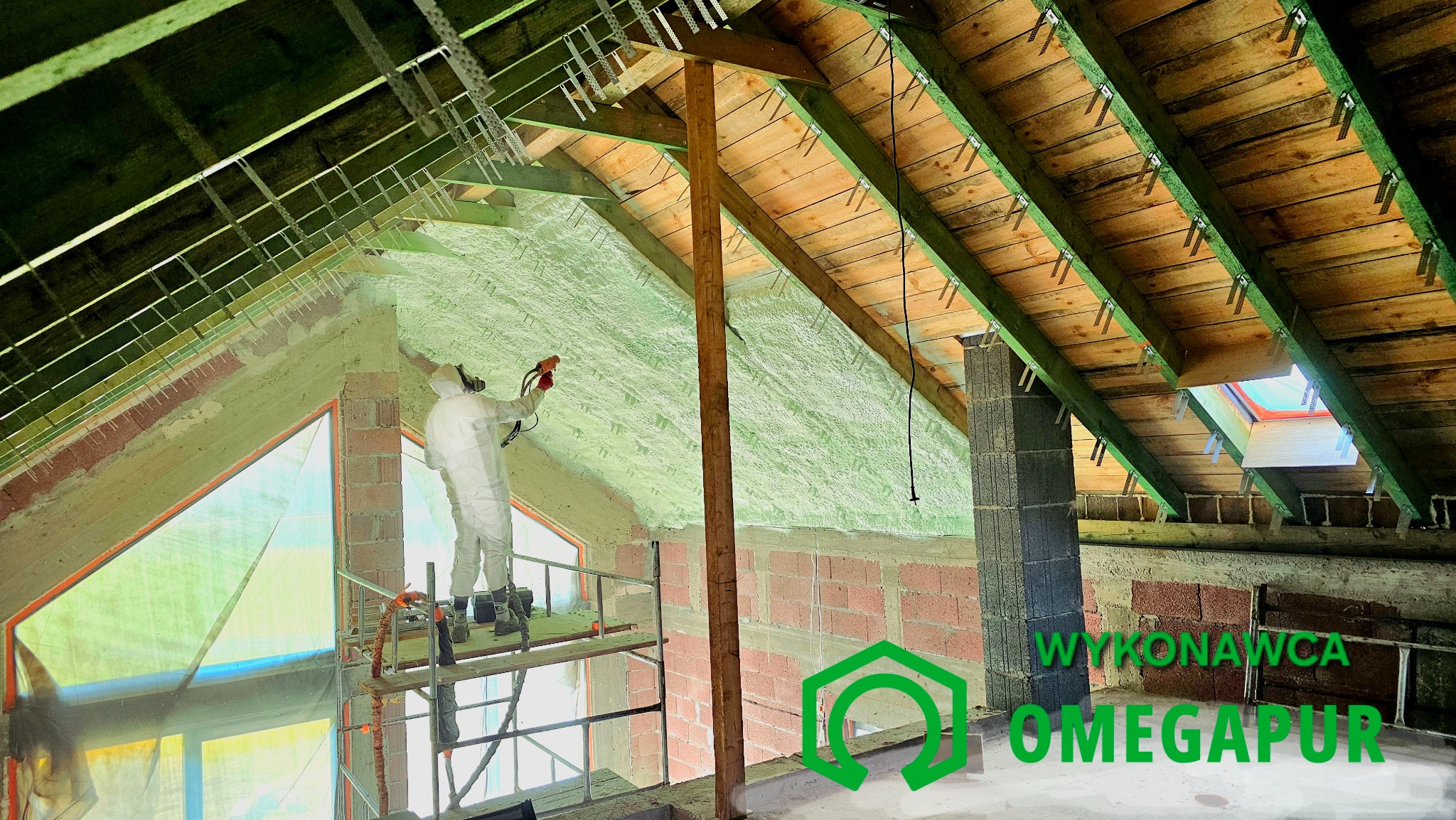 Pracownik w kombinezonie ochronnym aplikuje piankę izolacyjną na poddaszu, widoczne krokwie i elementy konstrukcyjne dachu, rusztowanie i logo firmy na pierwszym planie.