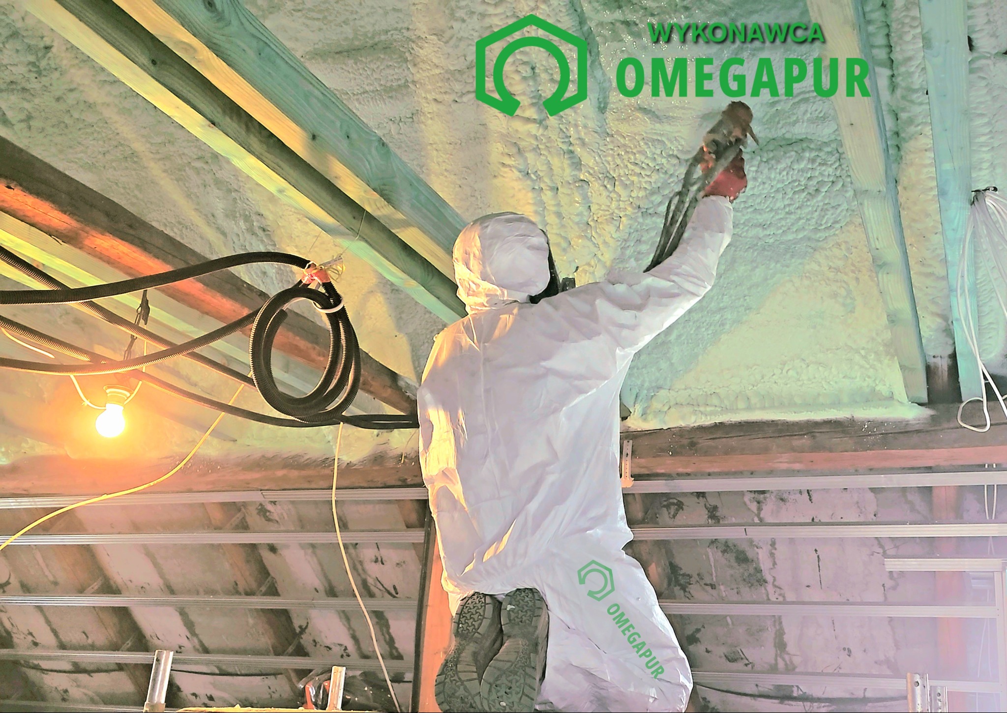 Pracownik w kombinezonie ochronnym aplikuje piankę na poddaszu, widoczne drewniane belki, instalacja elektryczna i logo firmy na kombinezonie.