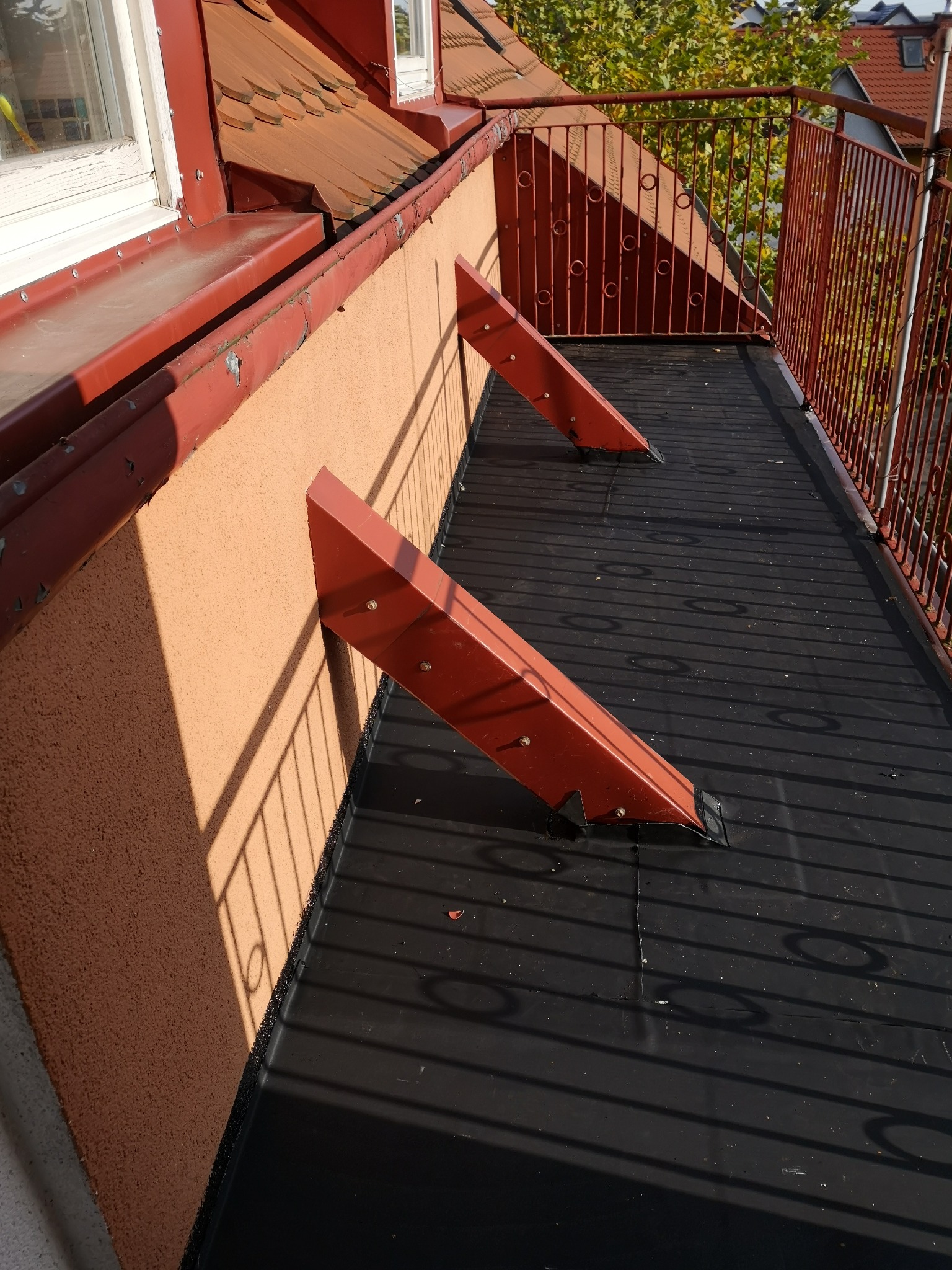 Balkon z nową, czarną izolacją przeciwwodną, wsparty na czerwonych, trójkątnych wspornikach przymocowanych do ściany budynku, z widoczną metalową balustradą i elementami dachu w tle.