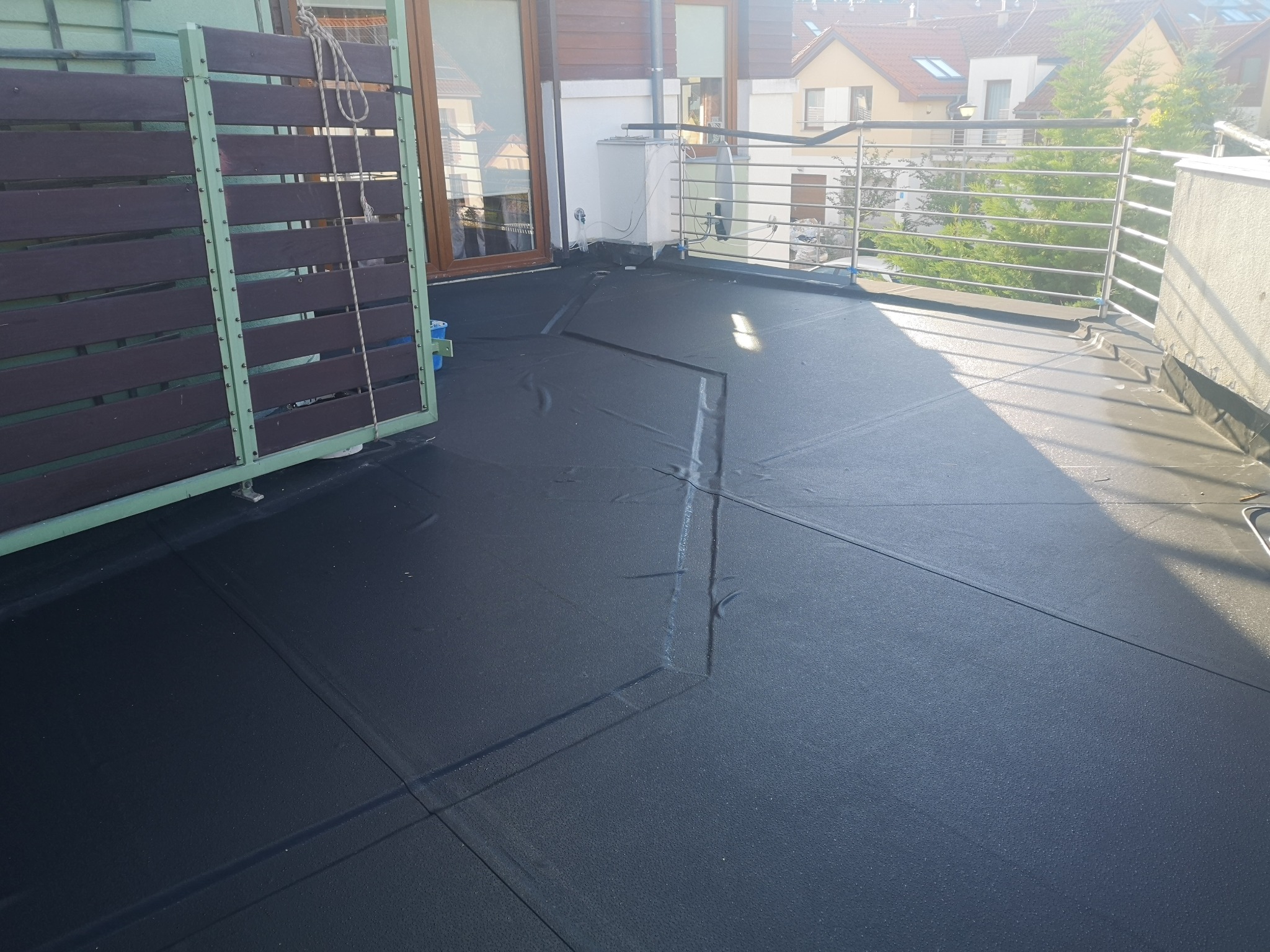 Czarna, gumowa izolacja przeciwwodna na tarasie, widoczne łączenia, fragment drewnianej osłony balkonu z zielonymi ramami, stalowa balustrada i budynki w tle.