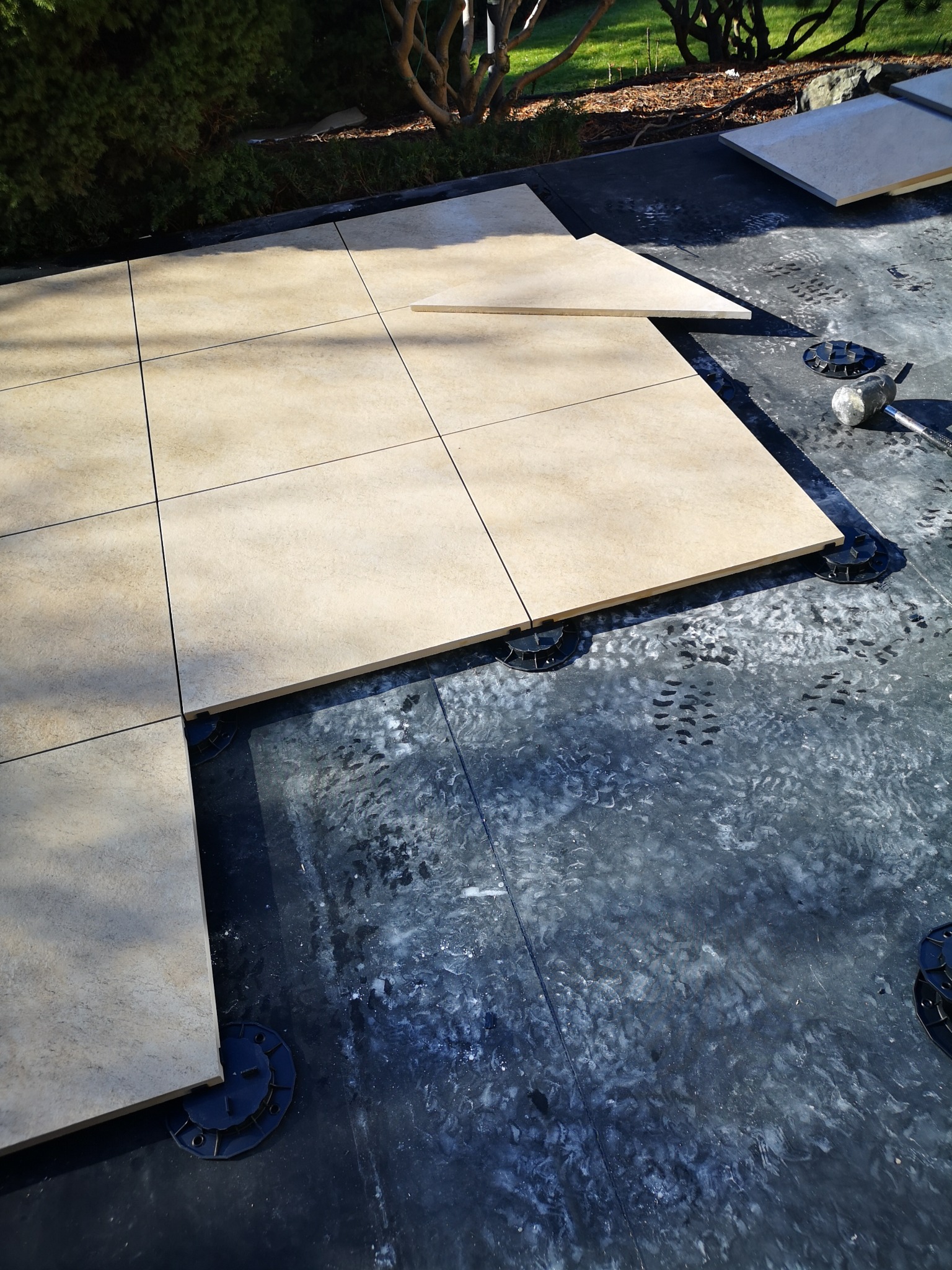 Układanie jasnobeżowych płyt na regulowanych wspornikach tarasowych na czarnej hydroizolacji, widoczne ślady robocze i gumowy młotek.