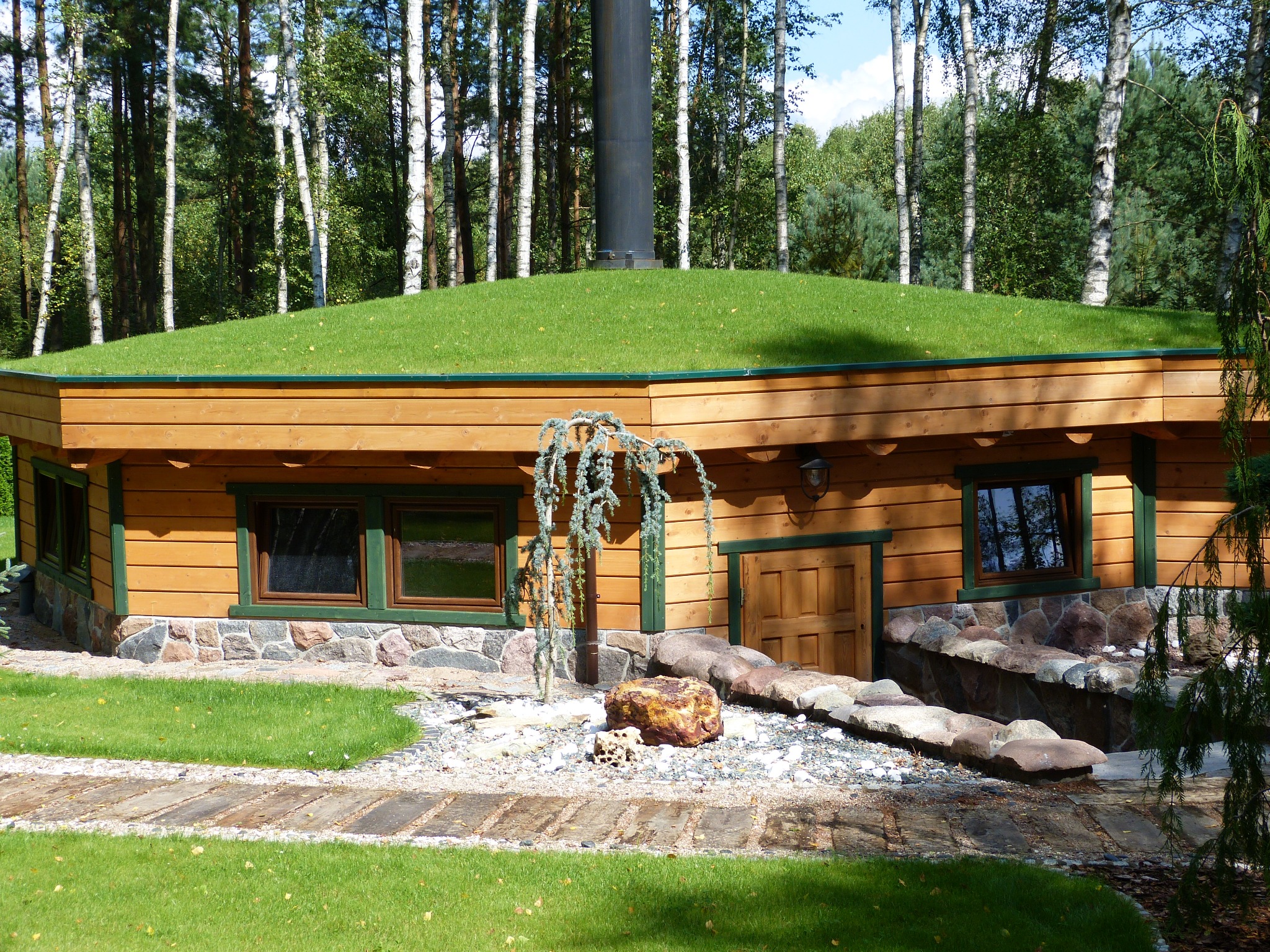 Drewniany budynek z zielonym dachem porośniętym trawą, z kominem, okna z zielonymi ramami, kamienna podmurówka i ścieżka z płyt.