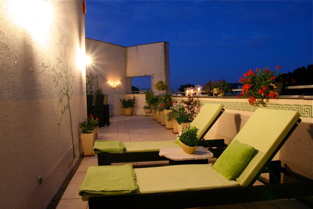 Umeblowany taras na dachu z dwoma leżakami, stolikiem, roślinami w donicach, oświetleniem ściennym i nocnym niebem w tle.