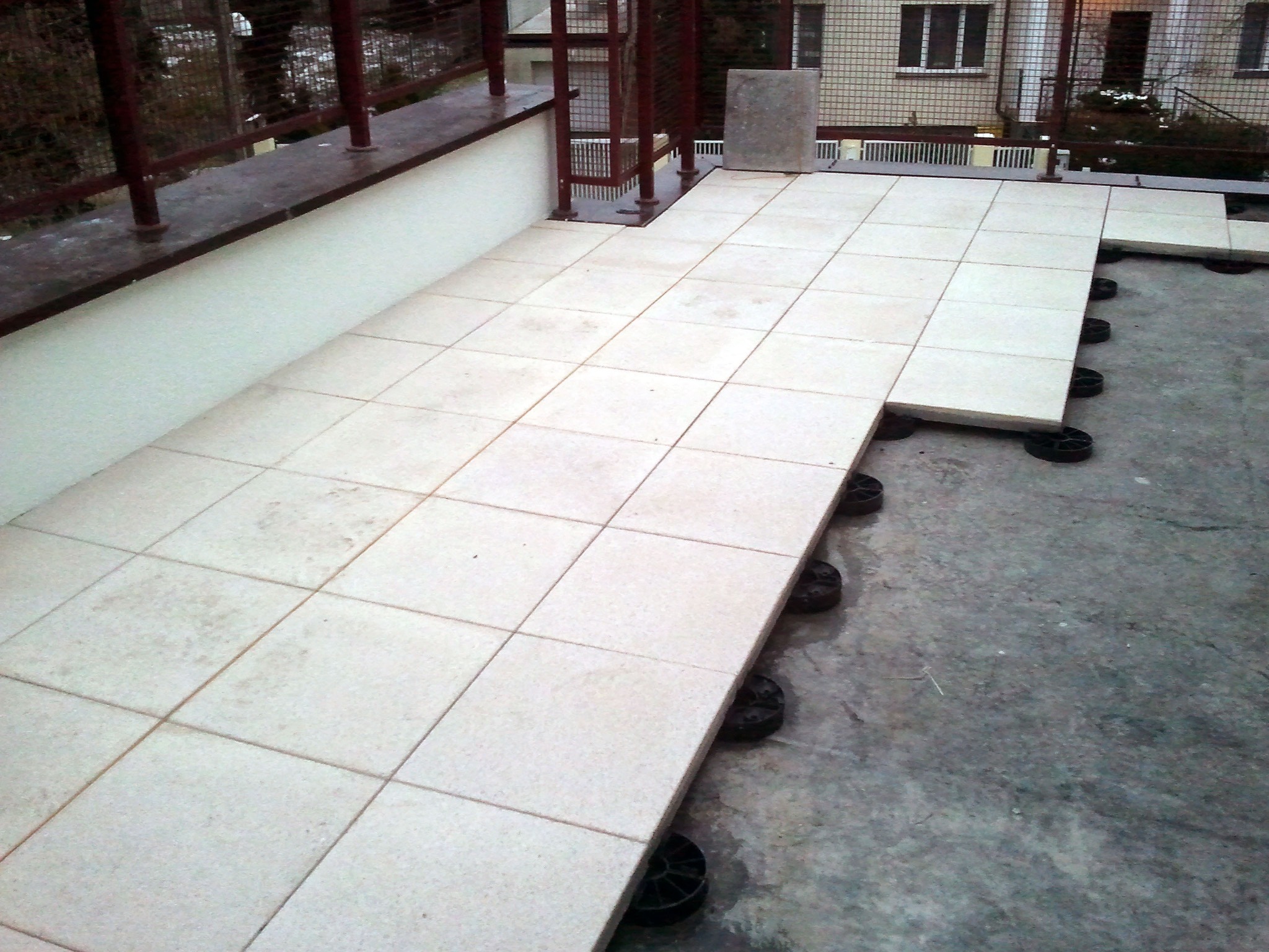 Montaż jasnych, kwadratowych płyt na regulowanych wspornikach na powierzchni balkonu, widoczne fragmenty metalowej balustrady i budynku w tle.