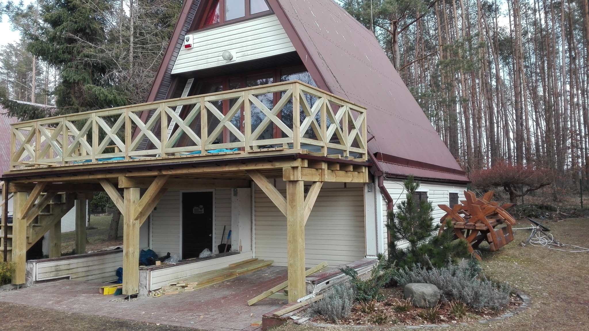 Dom w stylu A-frame z nowo wybudowanym, drewnianym tarasem z balustradą z krzyżowymi wzorami; widoczne narzędzia i materiały budowlane na ziemi.