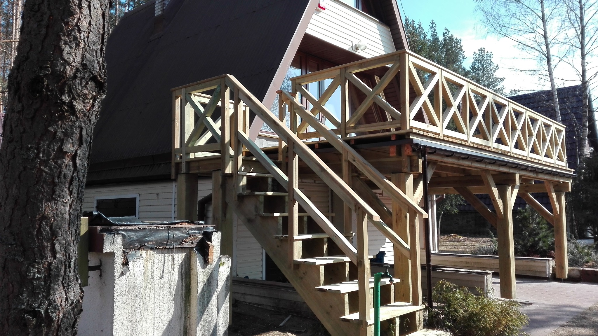 Drewniany taras z balustradą o geometrycznym wzorze krzyży, połączony schodami z ziemią, przylegający do domu z jasną elewacją i ciemnym dachem, widok z poziomu gruntu.