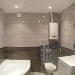 Wykończona łazienka z beżowymi płytkami, narożną wanną, umywalką z szafką, wiszącą toaletą i piecem gazowym z widoczną rurą odprowadzającą spaliny.