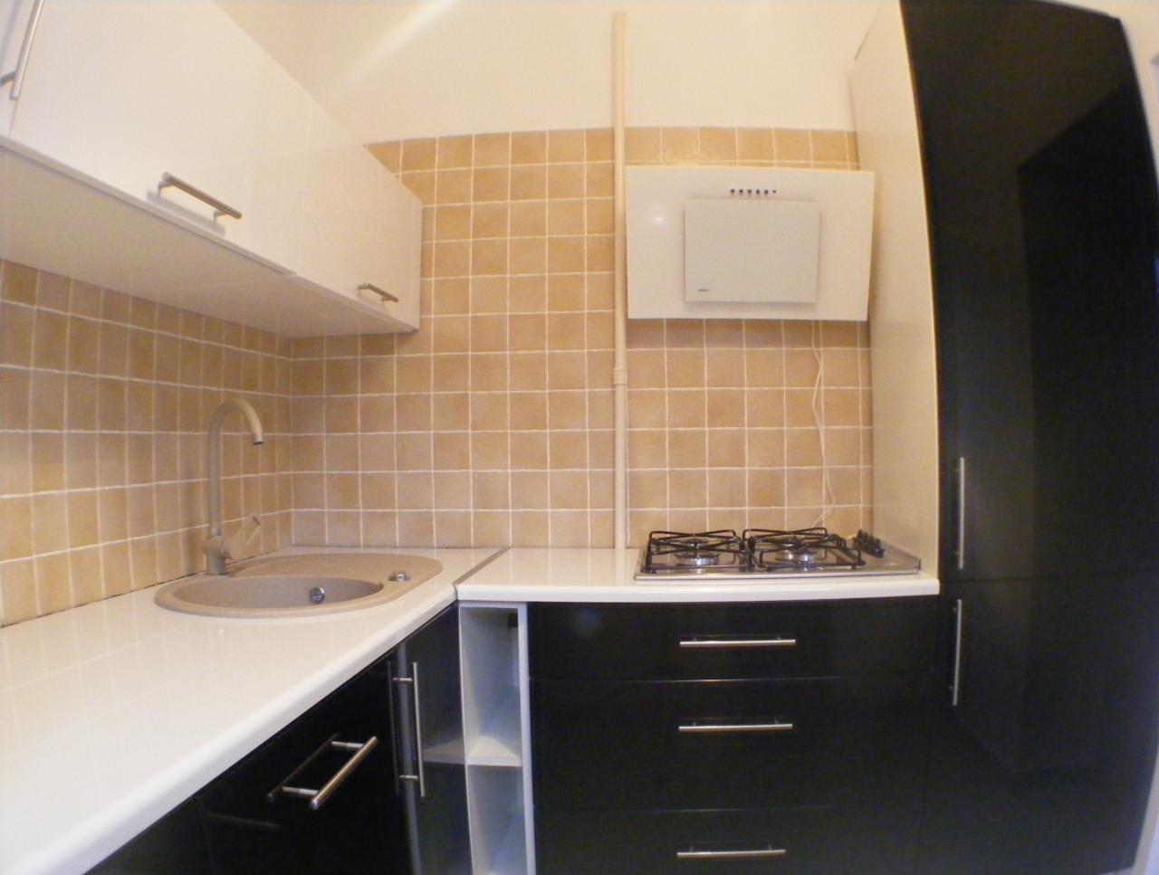 Wykończona kuchnia z beżowymi płytkami, czarnymi szafkami z poziomymi uchwytami, białym blatem i zlewem w odcieniach beżu, oraz biały okap nad płytą gazową.