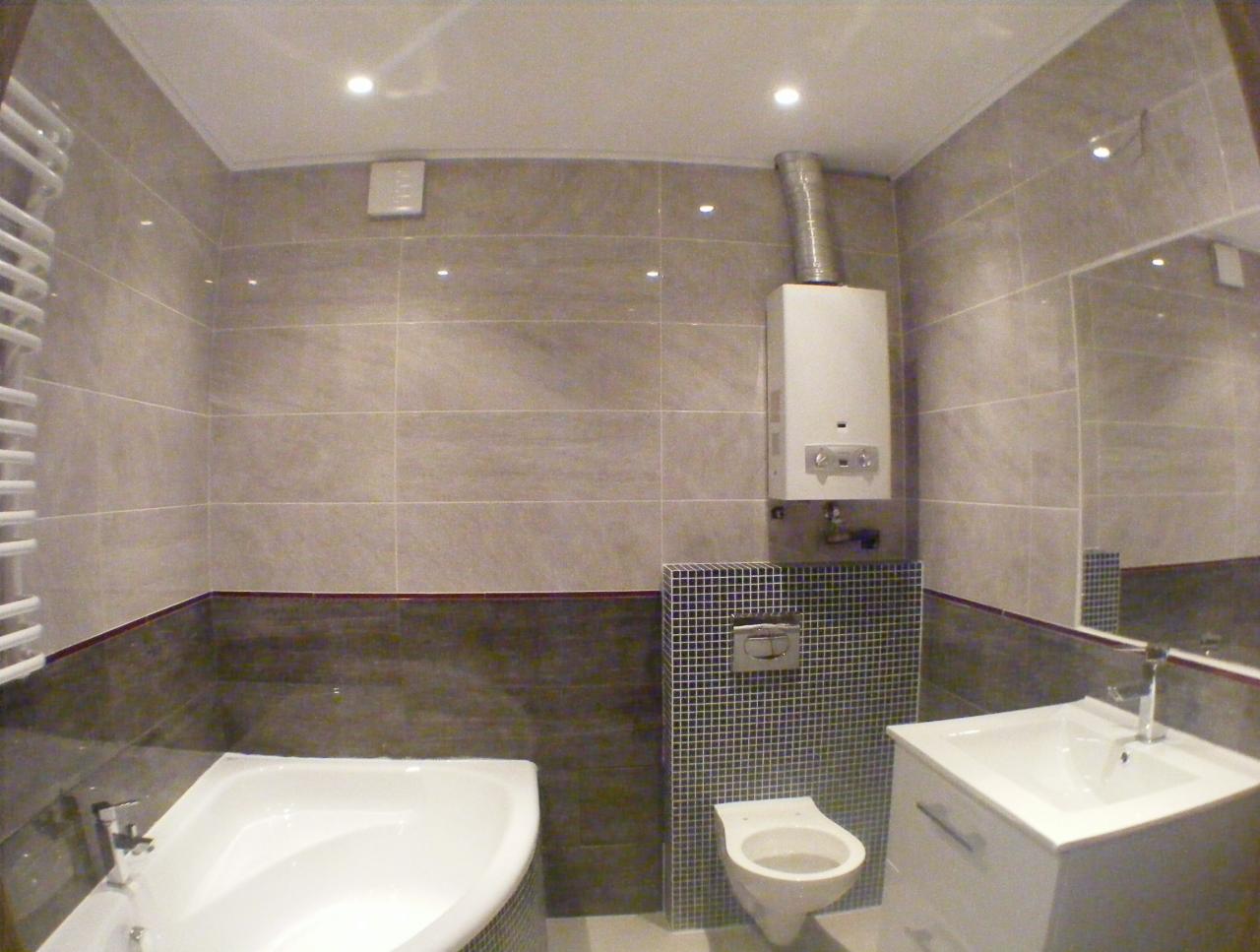 Wykończona łazienka z beżowymi płytkami, narożną wanną, umywalką z szafką, wiszącą toaletą i piecem gazowym z widoczną rurą odprowadzającą spaliny.