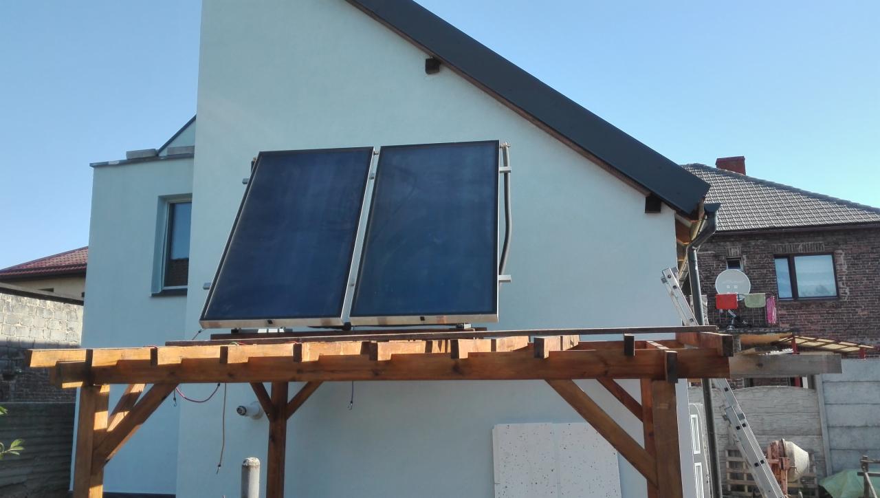 Dwa panele solarne zamontowane na drewnianej konstrukcji pergoli przy jasnym budynku, widoczna drabina i betoniarka w tle.