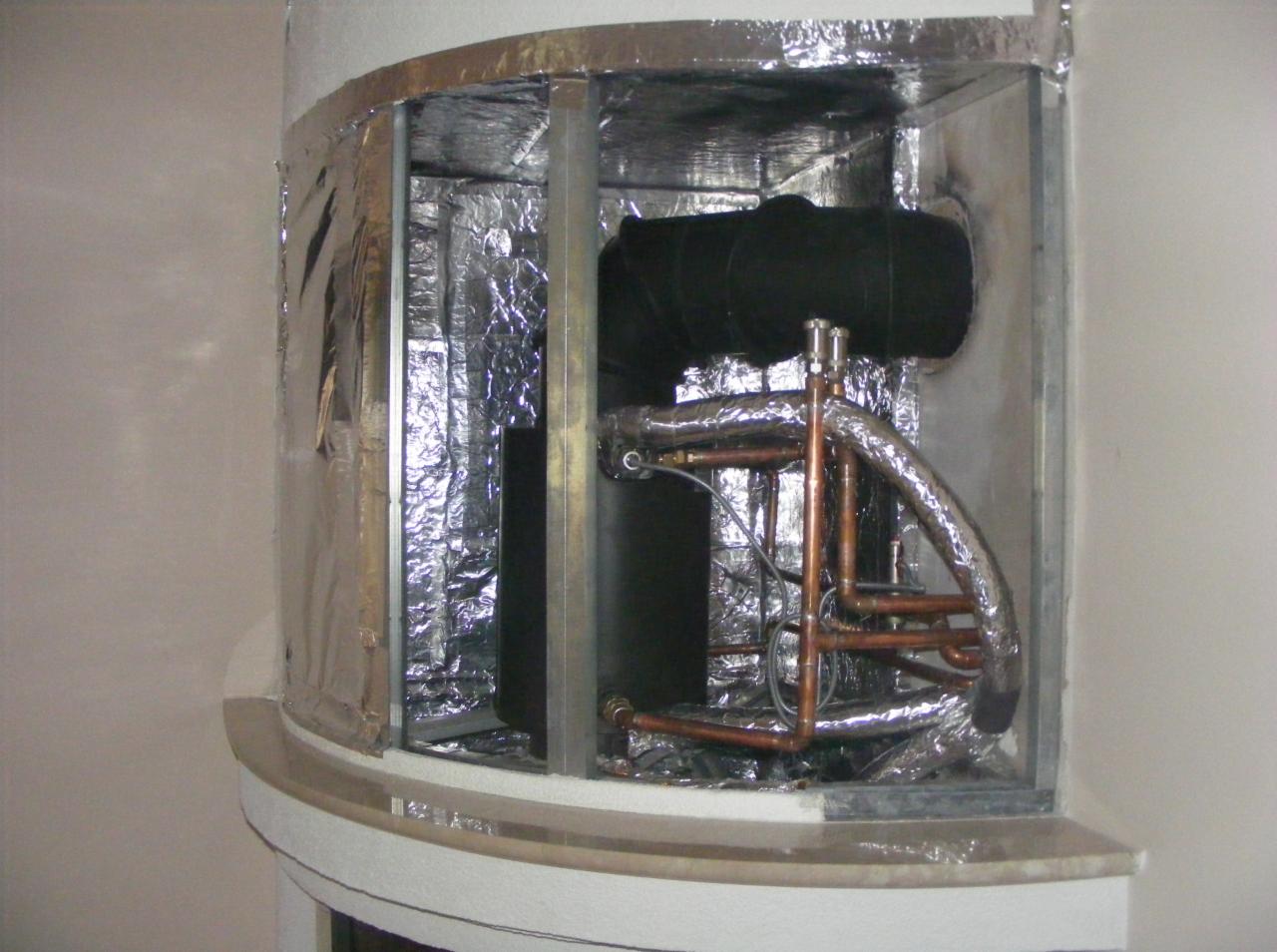 Wnętrze obudowy z izolacją termiczną, widoczne miedziane rury instalacji grzewczej i czarny element systemu, całość zamknięta w metalowej ramie.