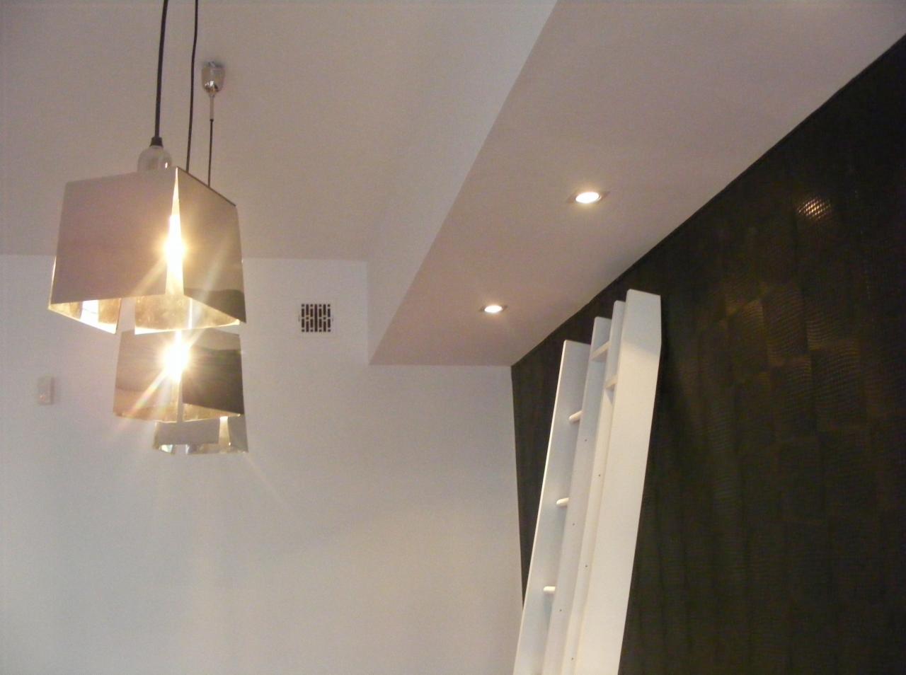 Nowoczesne wnętrze: geometryczna lampa wisząca z metalowymi panelami, biała drabina dekoracyjna oparta o ciemną ścianę z subtelnym wzorem, oraz wbudowane oświetlenie punktowe w suficie.