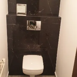 Zakończona realizacja: Toaleta z czarną marmurową glazurą na ścianie, podwieszana miska WC, szary dywanik na podłodze i biały grzejnik.
