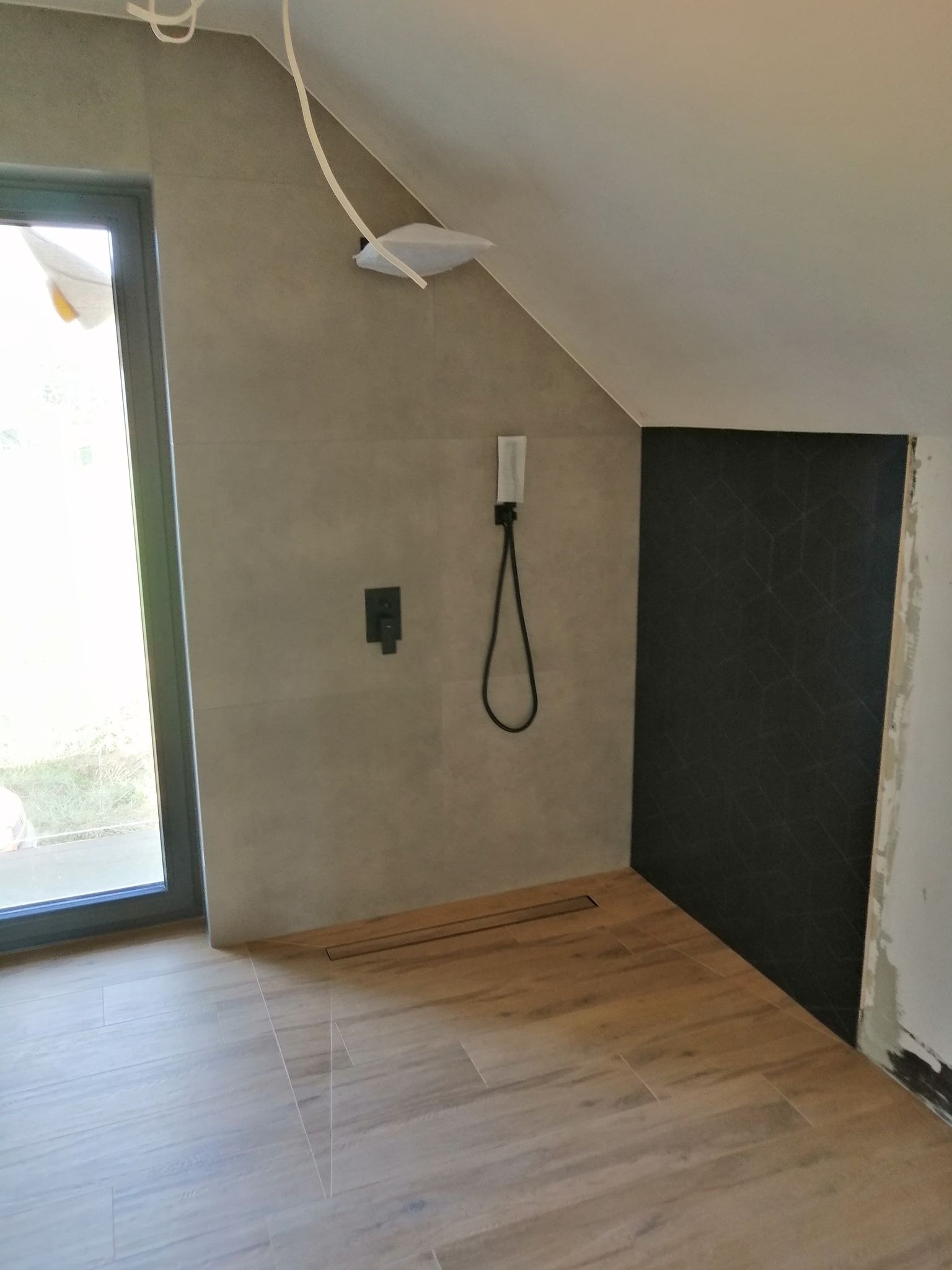 Wykończona łazienka na poddaszu z drewnopodobną podłogą, szarą glazurą ścienną i czarną ścianą z geometrycznym wzorem, widoczny odpływ liniowy i elementy instalacji podtynkowych.