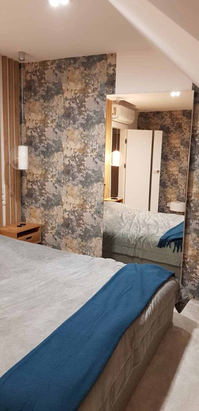 Sypialnia z łóżkiem, lampą wiszącą i tapetą imitującą beton, odbicie wnętrza w dużym lustrze stojącym.