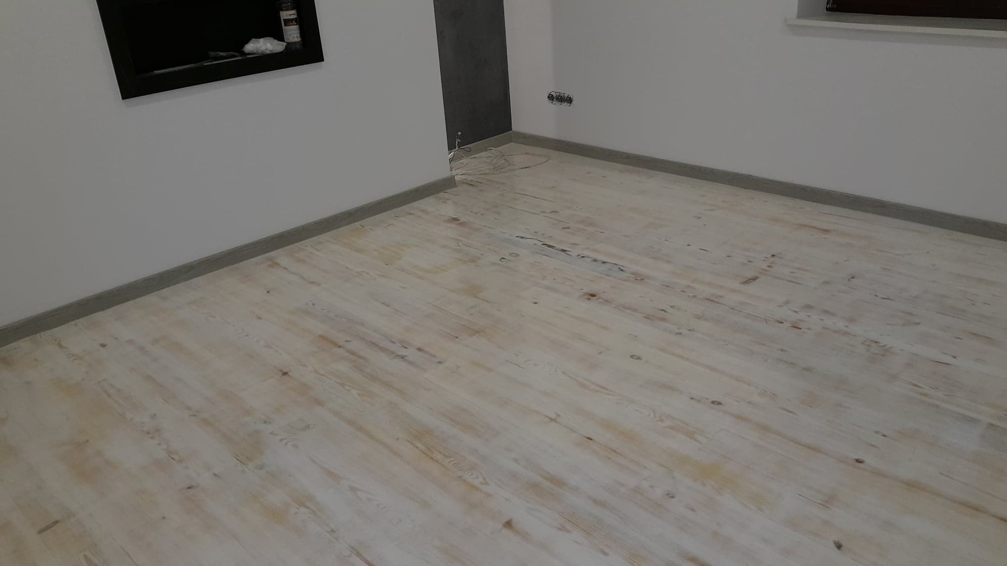 Jasna drewniana podłoga w trakcie odnawiania, widoczne ślady po szlifowaniu, surowe ściany z listwami przypodłogowymi, gniazdka elektryczne na ścianie.