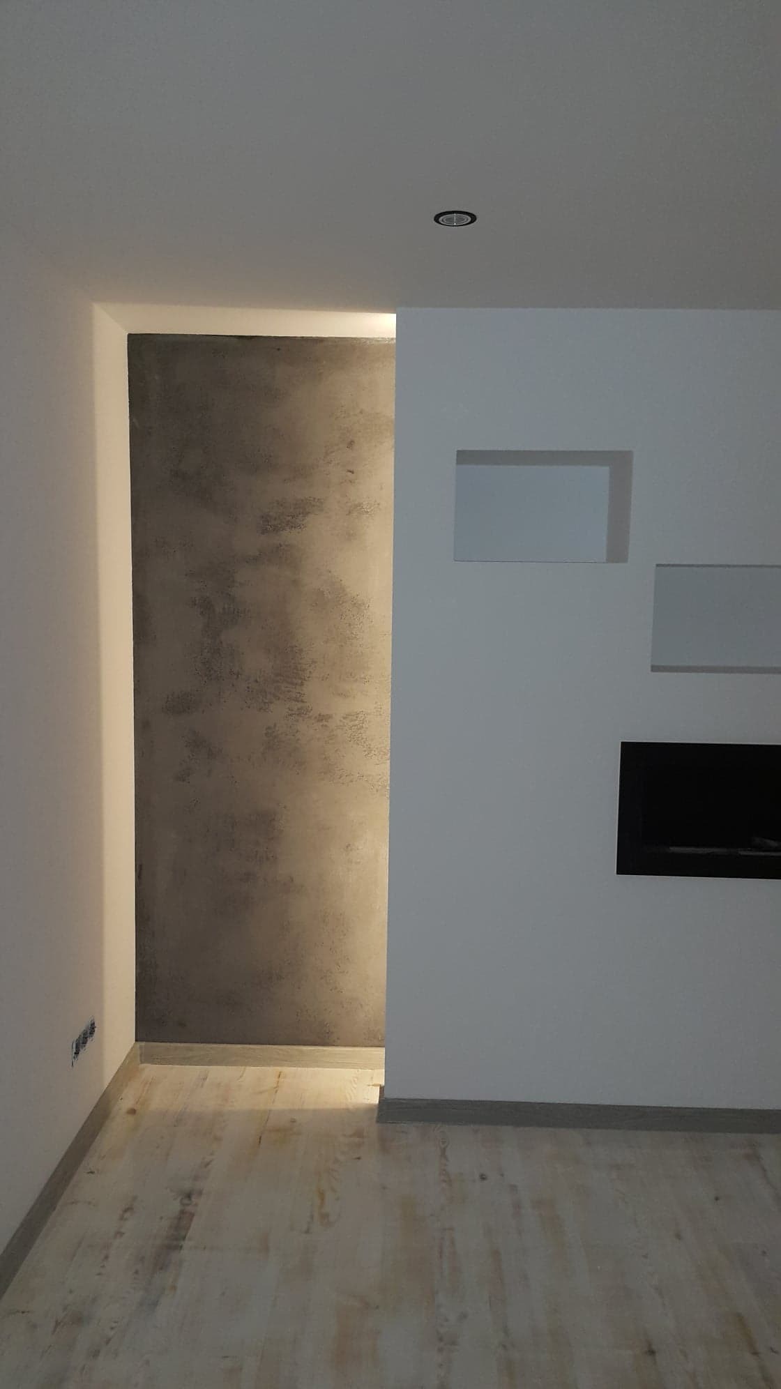 Fragment wnętrza po remoncie: ściana z dekoracyjną, szarą strukturą przypominającą beton architektoniczny, wnęki w białej ścianie oraz jasna drewniana podłoga.