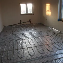 Rozłożone rury ogrzewania podłogowego na folii z siatką w surowym pomieszczeniu, widoczne okna i instalacje na ścianie.