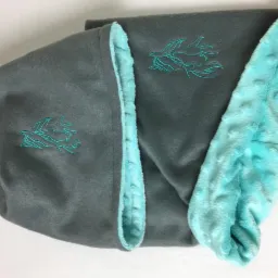 Szary kocyk z turkusową podszewką i haftowanym motywem roślinnym, złożony na białym tle.