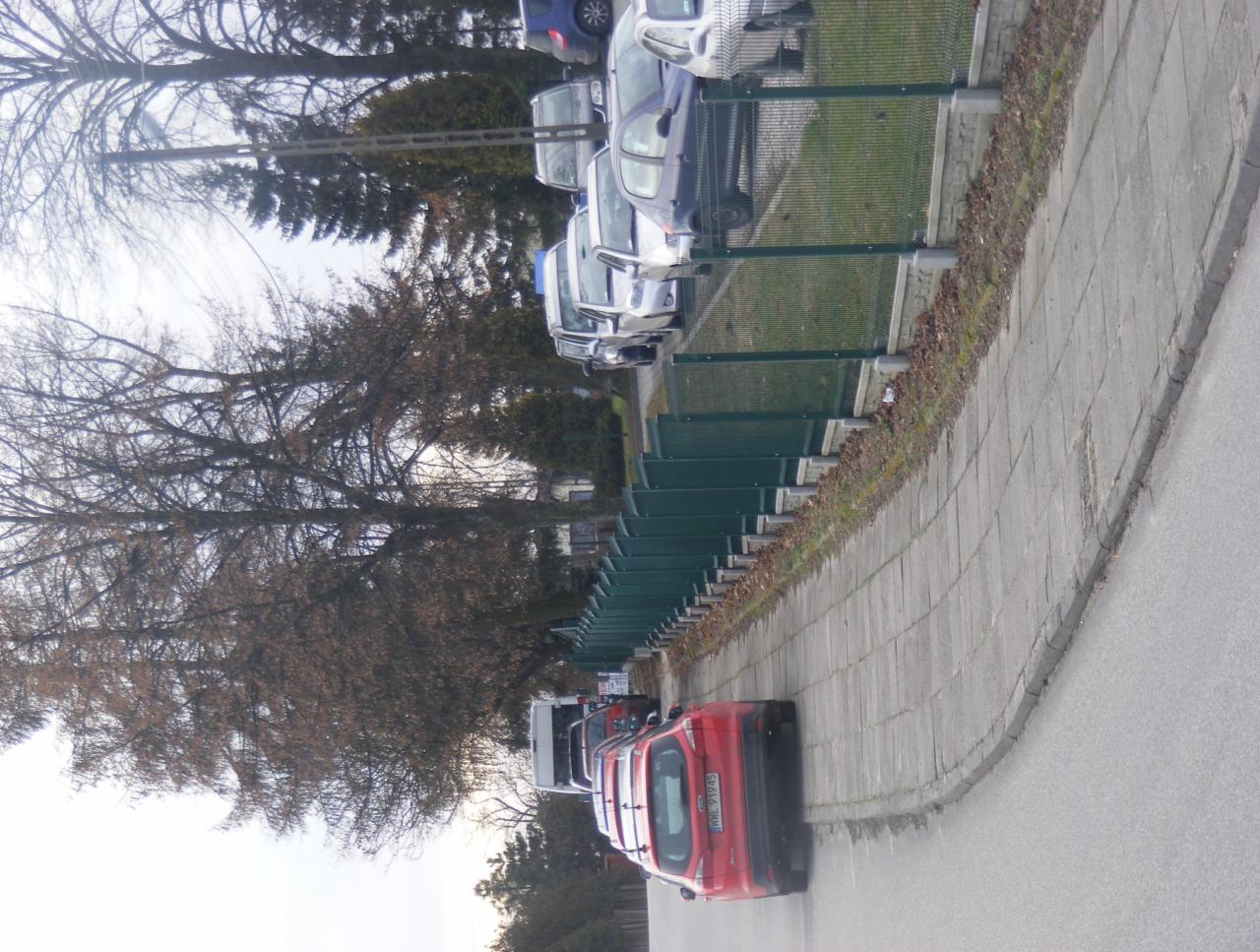 Szara kostka brukowa ułożona wzdłuż parkingu z zielonym ogrodzeniem, widok z poziomu chodnika w pochmurny dzień.