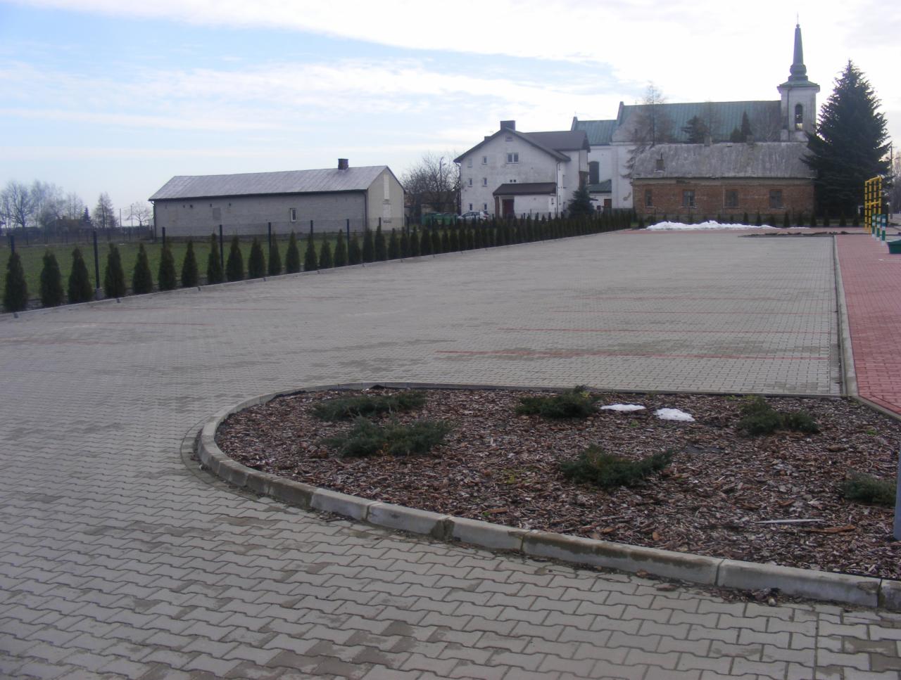 Rozległy parking z kostki brukowej w kształcie jodełki, obrzeża z tujami, w tle budynki, w tym kościół z wieżą.
