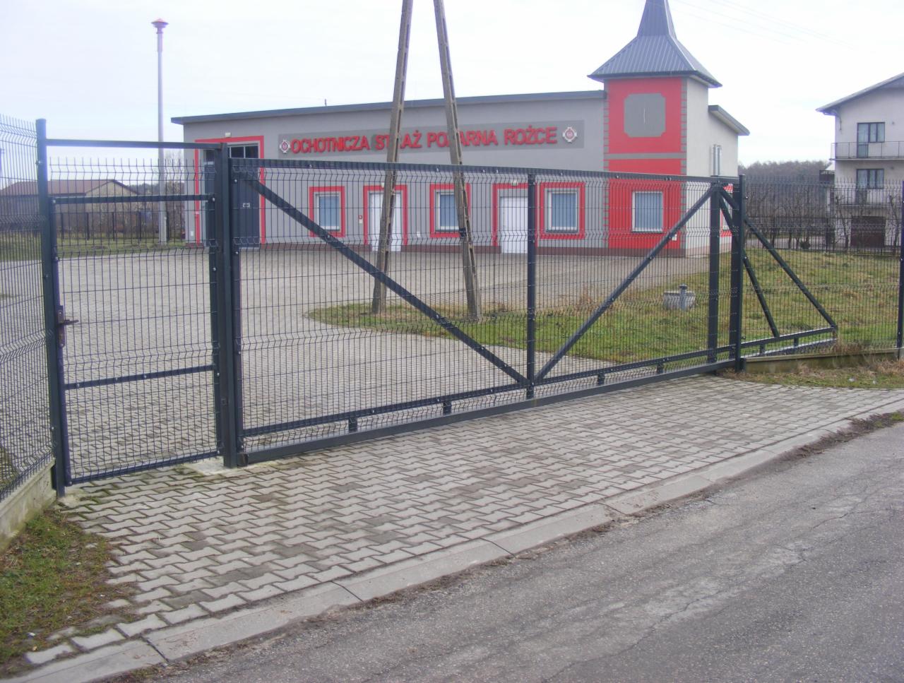 Ciemnoszara, metalowa brama przesuwna z siatki panelowej na posesji Ochotniczej Straży Pożarnej, widok z zewnątrz na kostkę brukową i budynek straży pożarnej w tle.