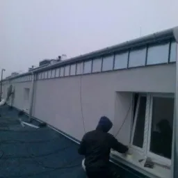 Pracownik w niebieskiej bluzie klęczy na pokryciu dachowym, uszczelniając okno na dachu budynku w pochmurny dzień.