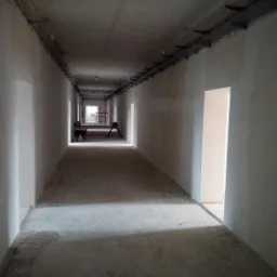 Długi korytarz w stanie surowym, świeżo po nałożeniu gładzi gipsowej, widoczne instalacje pod sufitem i drzwi bez ościeżnic.
