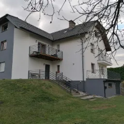 Dwukondygnacyjny dom z szarą i białą elewacją, balkonami z metalowymi balustradami, widoczny z poziomu trawnika w pochmurny dzień.