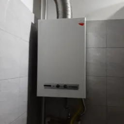 Biały, wiszący piec gazowy marki Termet z odprowadzeniem spalin w metalowym kominie, zamontowany na ścianie wyłożonej szarymi płytkami. Widoczne przyłącza gazowe i wodne pod urządzeniem.