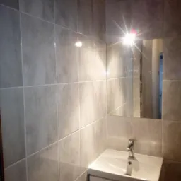 Wykończona łazienka z szarymi płytkami na ścianach, białą umywalką z szafką i prostokątnym lustrem, odbijającym światło lampy.