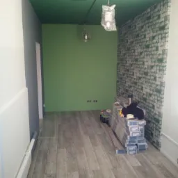 Wnętrze pokoju z ułożoną podłogą z paneli imitujących drewno, zielonymi ścianami i sufitem, oraz fragmentem ściany z tapetą imitującą cegłę. Na podłodze ułożone paczki paneli, widoczne gniazdka...